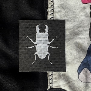 May include: A black fabric patch with a white silhouette of a stag beetle.