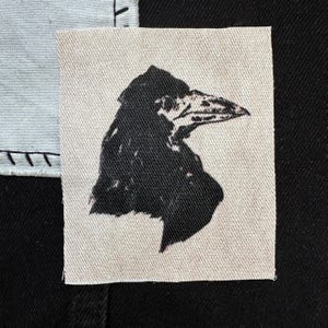 May include: A black and white embroidered patch of a crow's head with a sharp beak. The patch is on a white fabric background.