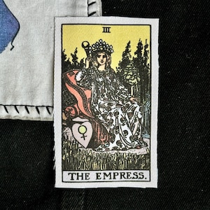 May include: A rectangular fabric patch featuring a tarot card illustration of The Empress. The design shows a crowned figure in a patterned gown holding a scepter, with a shield displaying a Venus symbol, set against a yellow and green background.