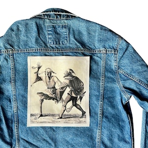May include: A blue denim jacket with a rectangular patch showing a black and white illustration of two figures in medieval clothing. The jacket has a classic design with a collar, button closure, and pockets.