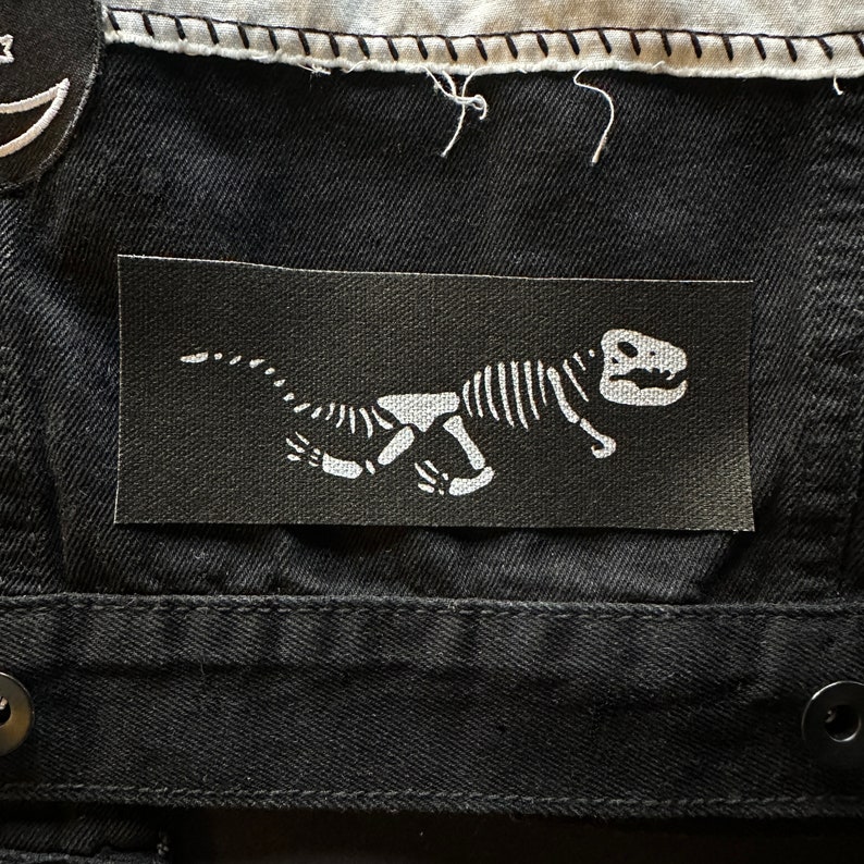 May include: Black fabric patch with a white dinosaur skeleton design. The dinosaur is facing right and has a long tail.