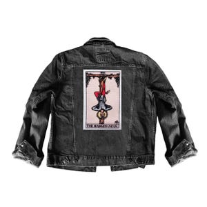 May include: Dark grey denim jacket with a rectangular patch displaying "The Hanged Man" tarot card. The card shows an upside-down figure, with text below. The jacket has a classic design with button fastenings and pockets.