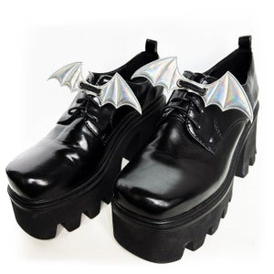 K&ouml;nnte beinhalten: Paar schwarze Plateauschuhe mit silbernen Fledermausfl&uuml;gel-Schuhschmuck, der an den Schn&uuml;rsenkeln befestigt ist.