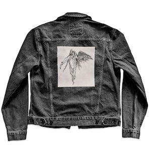 May include: A black denim jacket with a button-up front and a classic collar. The back features a square, off-white patch with a detailed black and white illustration of a winged figure. The jacket has two chest pockets and buttoned cuffs.