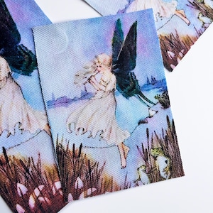 May include: A rectangular fabric patch featuring a printed illustration of a blonde figure with dark wings wearing a white dress, standing near tall brown reeds and green frogs against a blue sky background with a textured ribbed surface.