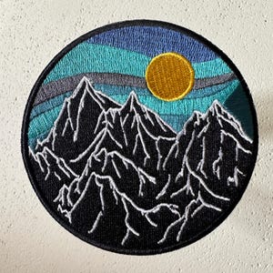 May include: Round embroidered patch featuring a mountain range silhouette in black with white detailing. The patch has a blue, teal, and gray sky with a yellow sun. The patch is approximately 3 inches in diameter.
