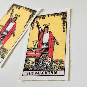 May include: Two rectangular fabric patches featuring the tarot card "The Magician." The card depicts a figure in a red robe with a candle, a table with objects, and the text "THE MAGICIAN" at the bottom. The background is yellow with floral accents.