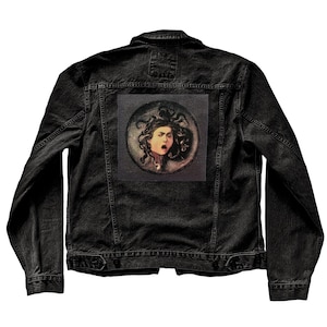 May include: Black denim jacket with a Medusa portrait patch on the back. The jacket has a classic design with button closures, pockets, and a collar. The patch features a detailed illustration of Medusa with her iconic snake hair.