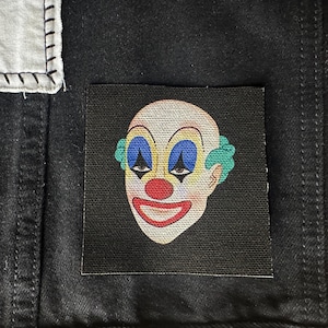 May include: A black fabric patch with a cartoon illustration of a clown's face. The clown has a white face with blue eyes, a red nose, and a red smile. The clown's hair is green.