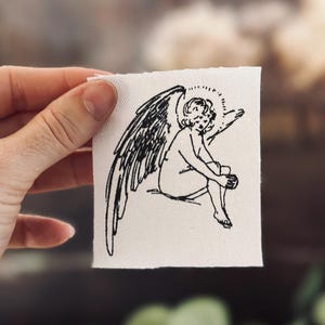 May include: A small, square, off-white fabric patch featuring a black line drawing of an angel with large wings and a halo. The angel is seated with one leg crossed over the other.