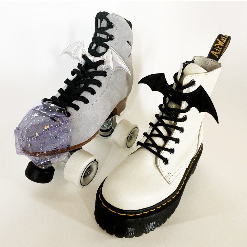 K&ouml;nnte beinhalten: Zwei Paar Rollschuhe mit Fledermausfl&uuml;gel-Anh&auml;ngern. Ein Paar ist wei&szlig; mit schwarzen Schn&uuml;rsenkeln und schwarzen Rollen. Das andere Paar ist grau mit schwarzen Schn&uuml;rsenkeln und schwarzen Rollen. Beide Paare haben eine glitzernde lila Zehenkappe.