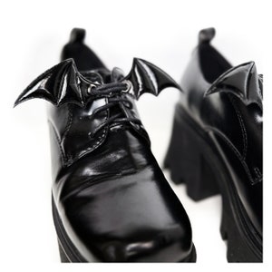 K&ouml;nnte beinhalten: Schwarze Plateauschuhe aus Leder mit Fledermausfl&uuml;gel-Schnallen. Die Schuhe haben einen Schn&uuml;rverschluss und eine dicke Plateau-Sohle.