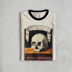 K&ouml;nnte beinhalten: Cremefarbenes Ringer-T-Shirt mit schwarzem Kragen und B&uuml;ndchen. Das Shirt zeigt eine detaillierte Illustration eines menschlichen Sch&auml;dels mit lateinischem Text auf einem Banner &uuml;ber und unter dem Sch&auml;del. Der Text enth&auml;lt "HIC DECOR HEC FORM-MNET" und "ANDREAS CA DI IO B P".