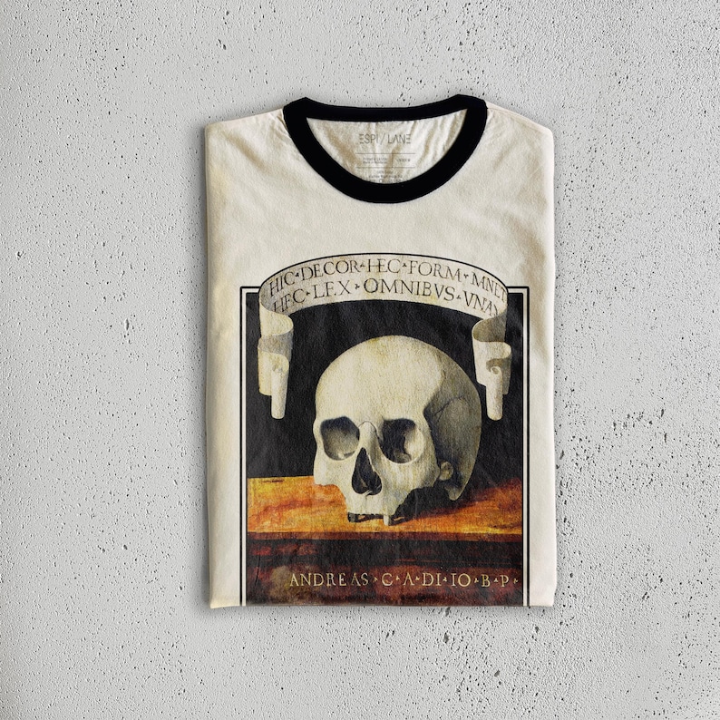 K&ouml;nnte beinhalten: Cremefarbenes Ringer-T-Shirt mit schwarzem Kragen und B&uuml;ndchen. Das Shirt zeigt eine detaillierte Illustration eines menschlichen Sch&auml;dels mit lateinischem Text auf einem Banner &uuml;ber und unter dem Sch&auml;del. Der Text enth&auml;lt "HIC DECOR HEC FORM-MNET" und "ANDREAS CA DI IO B P".
