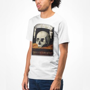 K&ouml;nnte beinhalten: Wei&szlig;es T-Shirt mit einer detaillierten Sch&auml;delillustration im klassischen Malstil, mit lateinischem Text. Das T-Shirt hat einen Rundhalsausschnitt und kurze &Auml;rmel.