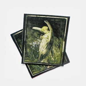 May include: Two rectangular patches featuring a vintage illustration of an angel in a garden. The angel is depicted in a flowing gown with large wings, surrounded by greenery and flowers. The patches have a black border and a textured appearance.