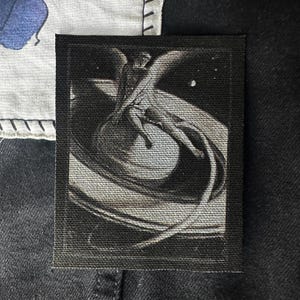 May include: Black and white embroidered patch featuring a stylized image of a person flying through space. The person is surrounded by a swirling, abstract design.