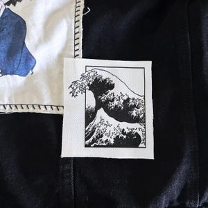 May include: A black and white patch featuring the iconic Japanese artwork, "The Great Wave off Kanagawa." The patch is rectangular with a black border and is sewn onto a black fabric. Another patch with a blue and white design is visible.