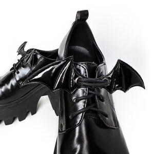 K&ouml;nnte beinhalten: Schwarze Plateauschuhe mit Fledermausfl&uuml;gel-Schn&uuml;rsenkeln. Die Schuhe haben eine klobige Plateau-Sohle und eine Schn&uuml;rung vorne.