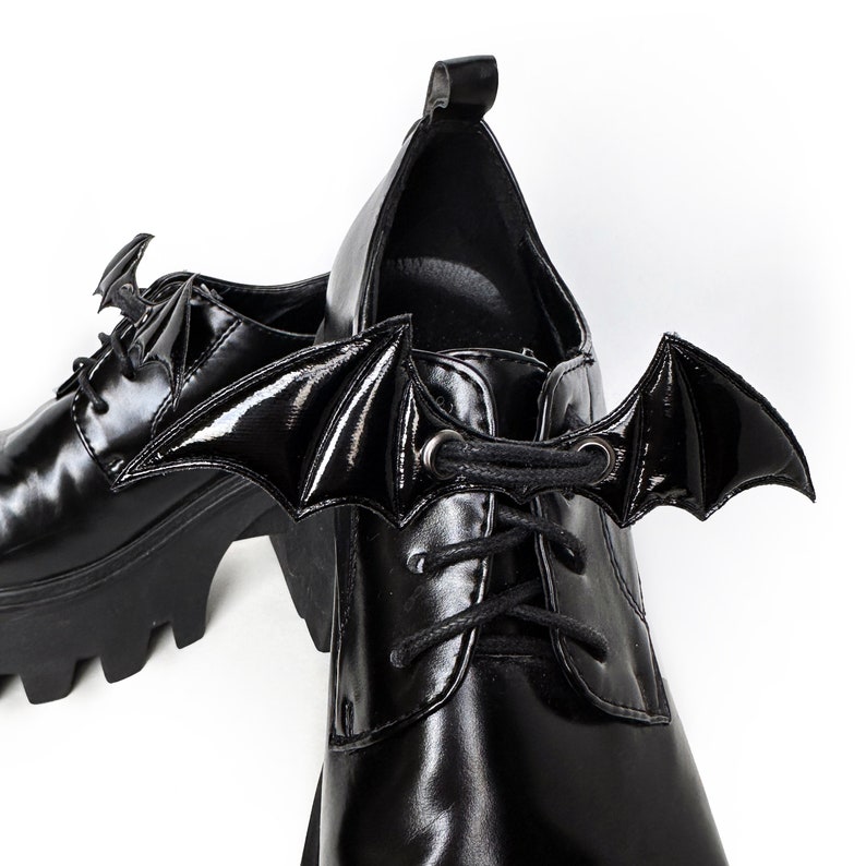 K&ouml;nnte beinhalten: Schwarze Plateauschuhe mit Fledermausfl&uuml;gel-Schn&uuml;rsenkeln. Die Schuhe haben eine klobige Plateau-Sohle und eine Schn&uuml;rung vorne.