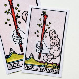 May include: Two rectangular fabric patches featuring the Ace of Wands tarot card design. Each patch displays a hand emerging from a cloud, holding a budding wooden wand, with a landscape below and the text ACE OF WANDS printed at the bottom.