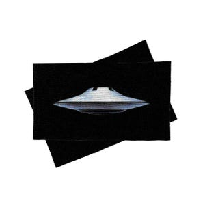 May include: A black fabric patch with a white flying saucer design. The saucer is in the center of the patch and is facing the viewer.