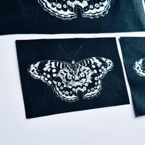 May include: A rectangular black fabric patch featuring a white screen-printed butterfly graphic with intricate wing patterns, including spots and lines. The design is centered on the fabric, showcasing a high-contrast monochrome aesthetic.
