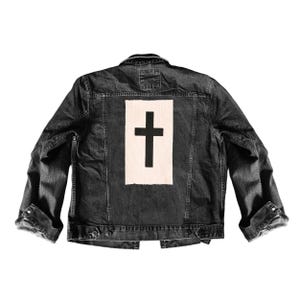 May include: A dark denim jacket with a black cross design on the back. The cross is centered on a white rectangular patch. The jacket has a classic collar, button closures, and long sleeves with buttoned cuffs. The jacket is a medium wash denim.