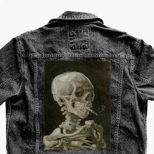 May include: Dark denim jacket featuring a painting of a skull smoking a cigarette. The artwork is a grayscale depiction of a skeleton with a cigarette in its mouth, set against a dark background. The jacket has a classic design with a collar and button closure.