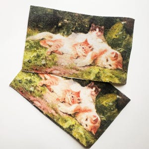 May include: Two rectangular fabric patches featuring a pastoral scene with cats. The image depicts several cats lounging in a grassy area, with a soft color palette of cream, peach, and green. The patches are slightly overlapping, with a white background.