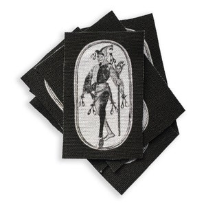 May include: A stack of black fabric patches with a white image of a jester holding a staff and wearing a hat with bells. The jester is standing in an oval frame.