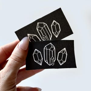 May include: Two black rectangular patches with white embroidered crystal designs. The crystals are geometric and stylized. The patches are held against a white background.
