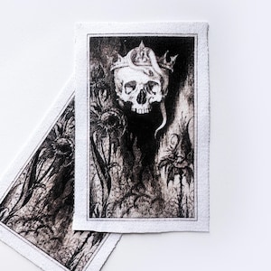May include: Two rectangular fabric patches with a black and white illustration of a skull wearing a crown, surrounded by stylized plants. The patches have a white border and are set against a white background.