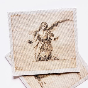 May include: Two square fabric patches with a sepia-toned illustration of an angel kneeling with outstretched arms and wings. The patches have a light beige border and are placed on a white background.