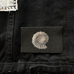 May include: A black fabric patch with a white embroidered ammonite fossil design.