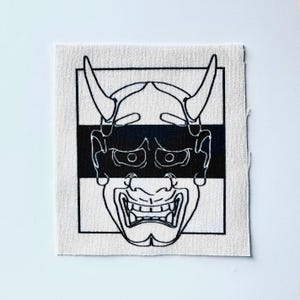 May include: A square, off-white fabric patch featuring a black and white illustration of a Japanese Oni mask. The mask has horns, a wide grin, and is framed by a black border and a horizontal black stripe.