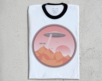 Könnte beinhalten: Weißes T-Shirt mit schwarzem Ringelkragen und einem Grafikdruck einer Wüstenlandschaft mit fliegenden Untertassen. Die Grafik befindet sich in einem Kreis mit einem rosa-orangenen Farbschema.
