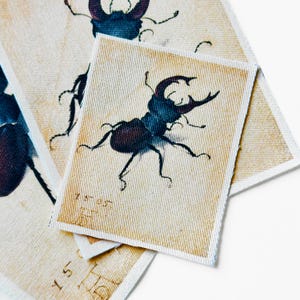 May include: Rectangular fabric patches with a vintage illustration of a beetle. The beetle is depicted in shades of blue and purple, with intricate details. The background is a beige color, and the patches have a textured appearance. Some patches have handwritten numbers.
