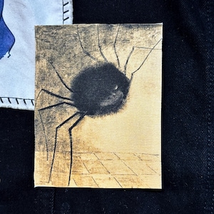 May include: A small, rectangular canvas patch featuring a black spider illustration. The spider has a fuzzy body and long, thin legs. The background is a light tan color with a grid pattern at the bottom.