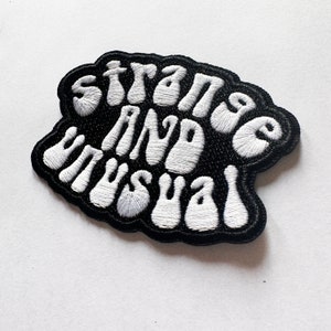 May include: Black embroidered patch with white lettering that reads "Strange and Unusual".
