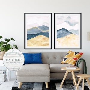 May include: Two framed watercolour mountain prints in black frames. The artwork features blue and gold hues. The image includes the text "Starchhouse" and "Mix & Match each sold separately". The prints are displayed above a grey sofa.