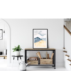 May include: A framed watercolour print depicting a mountain scene in blue and gold hues. The artwork is displayed on a wooden console table with a black metal frame. The room features a white wall, a small side table with a plant, and a staircase.