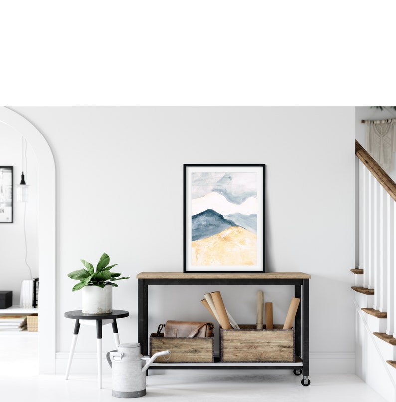 May include: A framed watercolour print depicting a mountain scene in blue and gold hues. The artwork is displayed on a wooden console table with a black metal frame. The room features a white wall, a small side table with a plant, and a staircase.