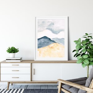 May include: A framed watercolour painting depicting abstract mountains in blue and gold hues. The artwork is displayed on a white cabinet with wooden detailing, alongside a small potted plant and a modern armchair. The room has a minimalist design.