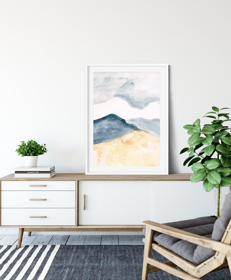 May include: A framed watercolour painting depicting abstract mountains in blue and gold hues. The artwork is displayed on a white cabinet with wooden detailing, alongside a small potted plant and a modern armchair. The room has a minimalist design.