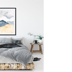 May include: A framed watercolour painting of a mountain landscape in shades of blue and gold hangs above a bed with grey bedding. The room has a minimalist design with a wooden side table and a desk lamp.
