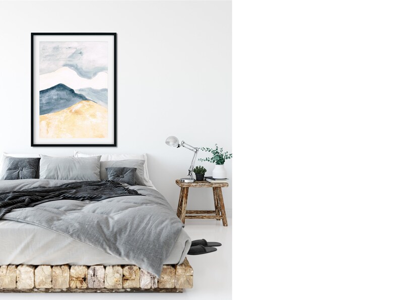 May include: A framed watercolour painting of a mountain landscape in shades of blue and gold hangs above a bed with grey bedding. The room has a minimalist design with a wooden side table and a desk lamp.