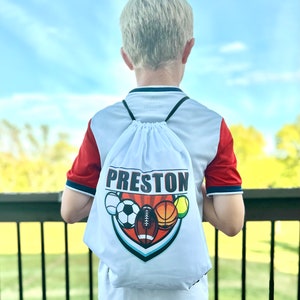 May include: White drawstring bag with a colorful graphic of sports balls and the name Preston printed in red.