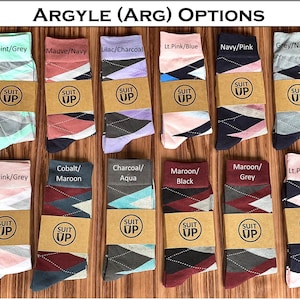 May include: A collection of argyle socks in various color combinations, including mint/grey, mauve/navy, and lilac/charcoal. Each pair is wrapped with a brown paper band featuring the text "SUIT UP". The socks are displayed on a wooden surface.
