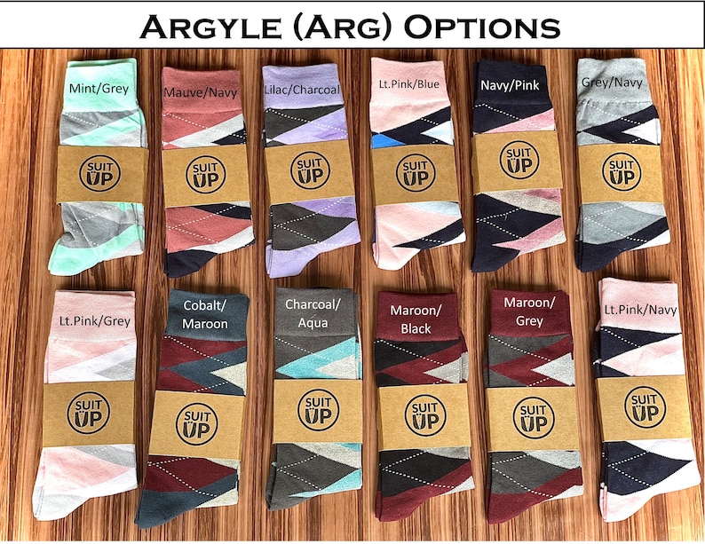 May include: A collection of argyle socks in various color combinations, including mint/grey, mauve/navy, and lilac/charcoal. Each pair is wrapped with a brown paper band featuring the text "SUIT UP". The socks are displayed on a wooden surface.